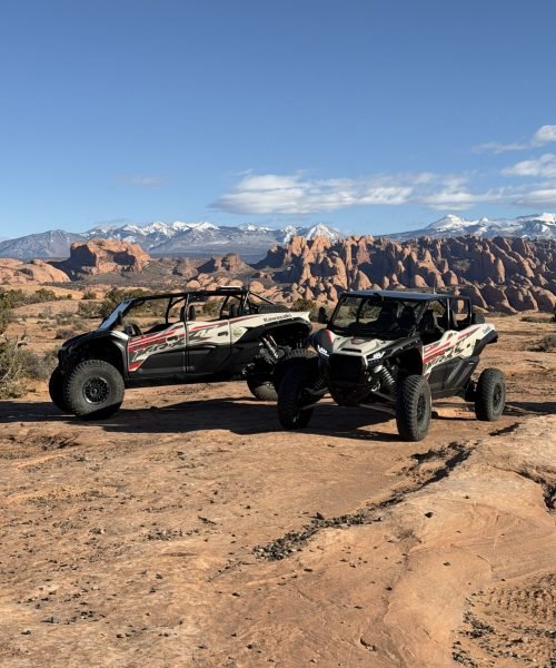Two all-terrain vehicles parked on a desert with red rock formations and snow-capped mountains under a clear blue sky.